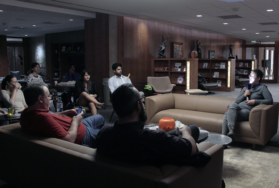 A group of people sit on couches in a dimly lit room. A person with a flashlight on their face shares a research horror story with the group.